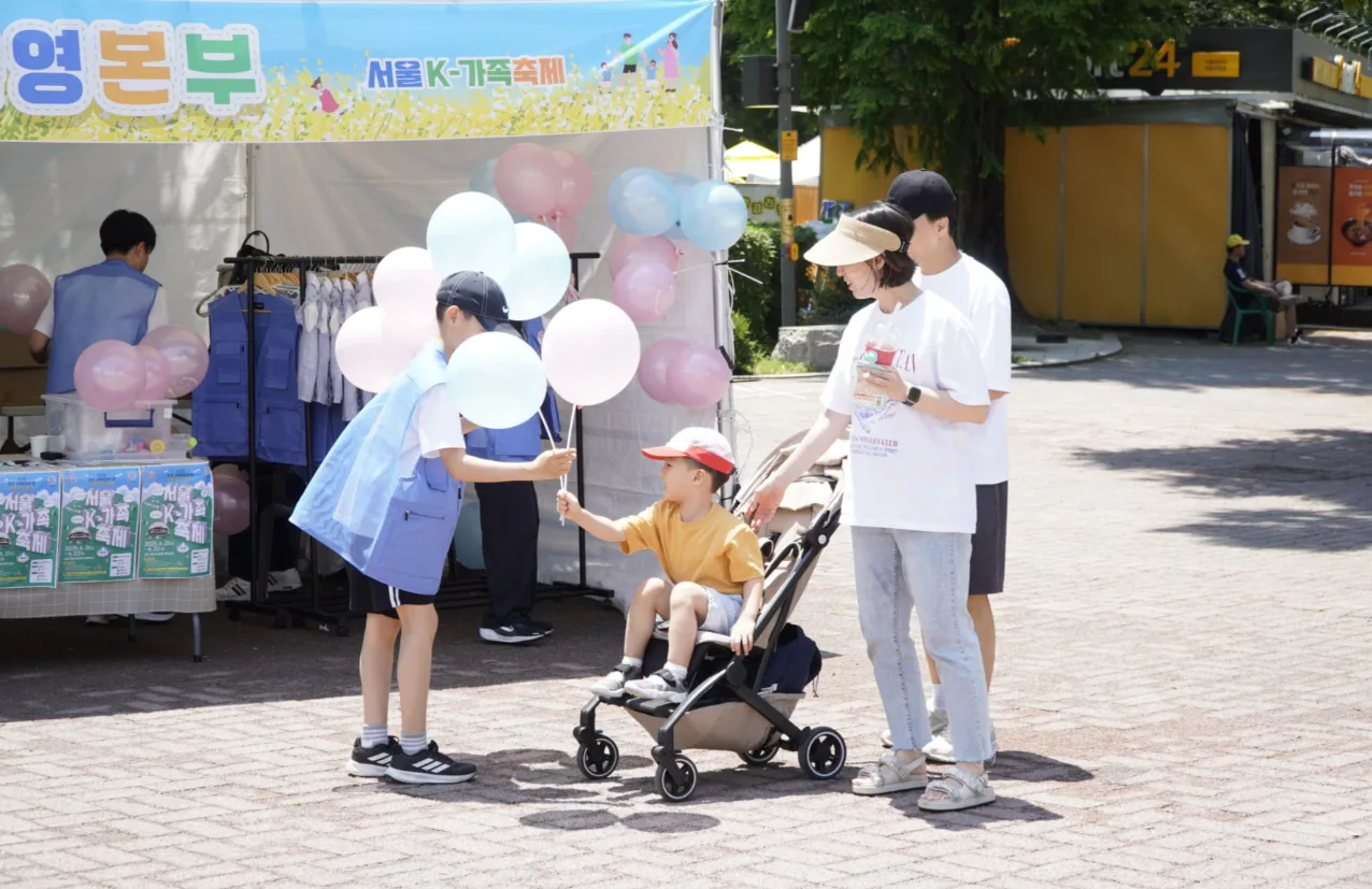 2025년 서울 K-가족축제에서 파란 조끼를 입은 어린이 봉사자가 유모차에 탄 아이에게 풍선을 건네고 부모가 곁에서 지켜보는 모습