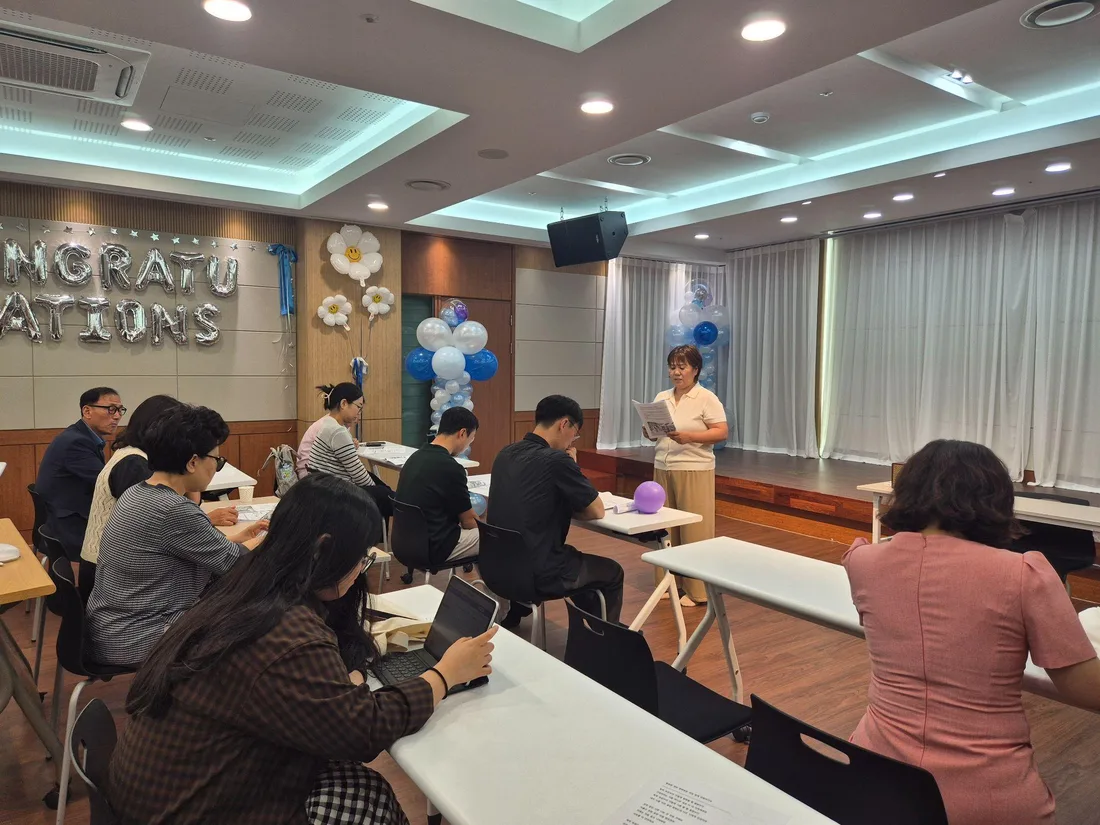 풍선으로 꾸민 강당에서 진행자가 자료를 읽으며 설명하고, 참석한 부모들이 책상에 앉아 듣고 있는 모습