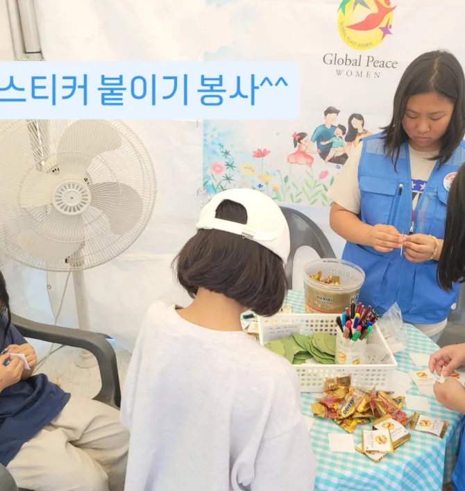 파란 조끼를 입은 청소년 봉사자가 부스 탁자에서 체험 스티커를 준비하고 어린이 참가자들이 둘러앉은 모습. 사진 위에 ‘스티커 붙이기 봉사’라는 글자가 얹혀 있다