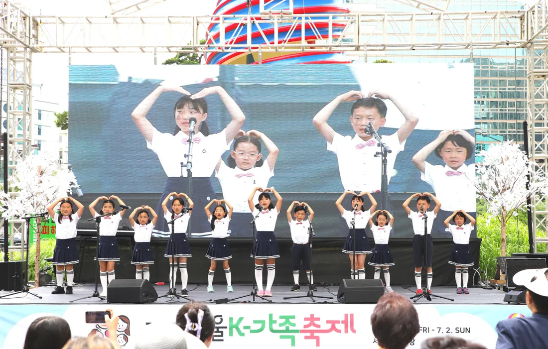 흰 셔츠와 남색 치마·바지를 입은 어린이 중창단이 머리 위로 하트를 만들며 노래하고, 뒤편 대형 화면에 아이들 모습이 비치는 서울 K-가족축제 무대