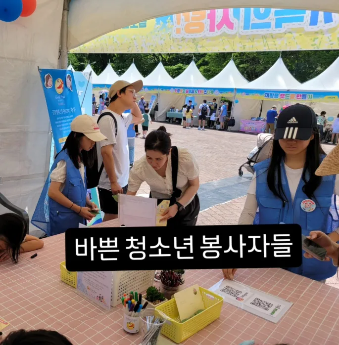 흰 천막 부스가 늘어선 축제장에서 파란 조끼를 입은 청소년 봉사자들이 체험 탁자 앞의 시민을 안내하는 모습. 사진 위에 ‘바쁜 청소년 봉사자들’이라는 글자가 얹혀 있다